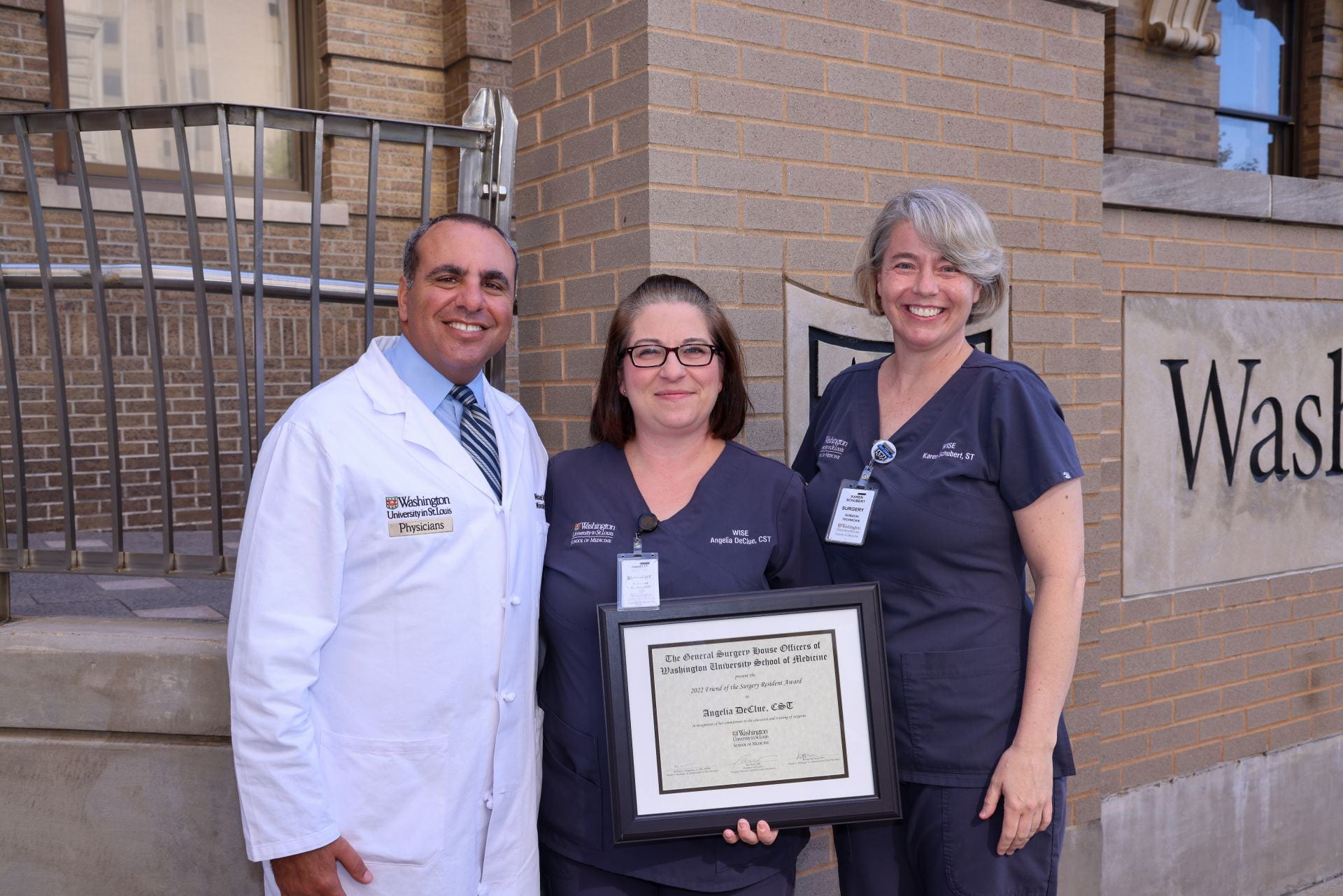 Michael Awad in white coat with WISE staff in blue scrubs holding award in front of brick exterior of School of Medicine building.