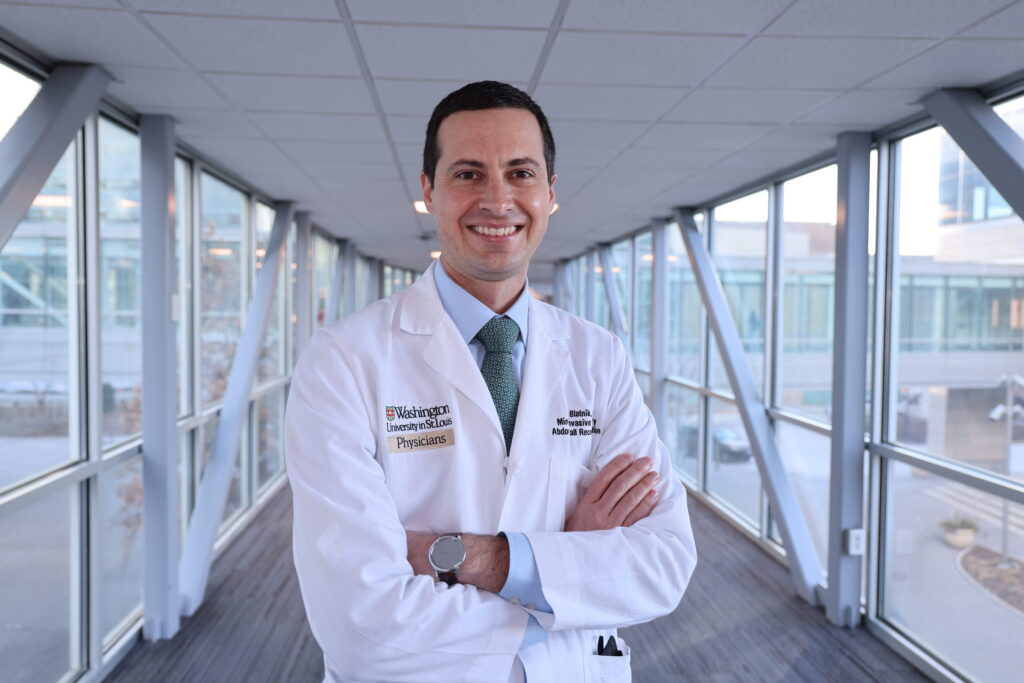 Dr. Blatnik smiling and wearing white coat standing in enclosed walkway with arms crossed.